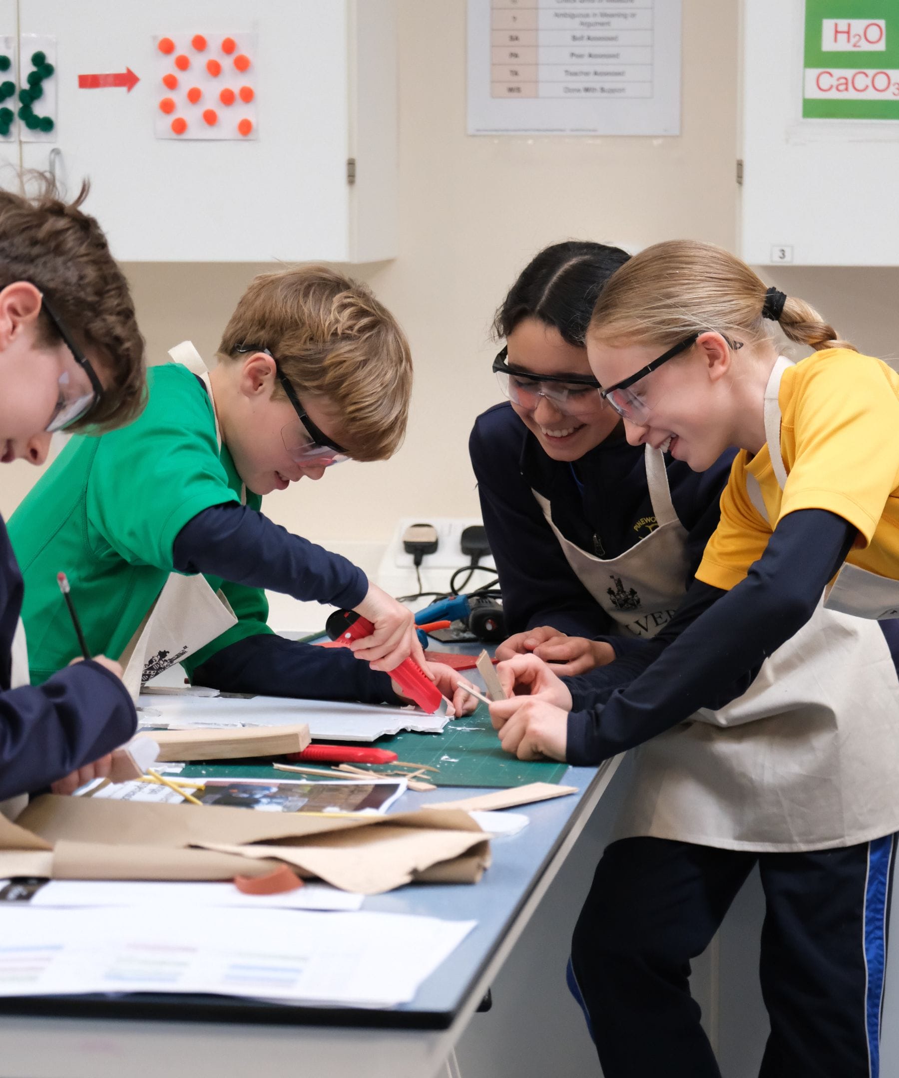 Four pupils in colourful tops wearing safety goggles whilst smiling between themselves as they cut board